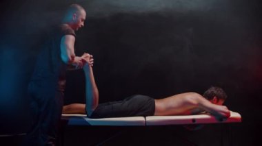 Karanlık stüdyoda kiropraktik terapi. Müvekkilleriyle birlikte çalışan bir terapist. Bacağını büküyor ve çabalayarak bastırıyor.