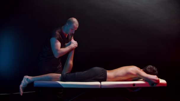Хіропрактична терапія в темній студії - терапевт, що вигинає ногу свого клієнта, коли вагається лежати на животі — стокове відео