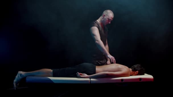 Chiropraxis terápia a sötét stúdióban - férfi ügyfél fekszik a kanapén, és a terapeutája nyomja a pengéit — Stock videók