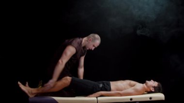 Karanlık stüdyoda kiropraktik terapi. Müvekkilinin bacaklarıyla çalışan bir terapist. Bacaklarını vücuduna bastırıyor.