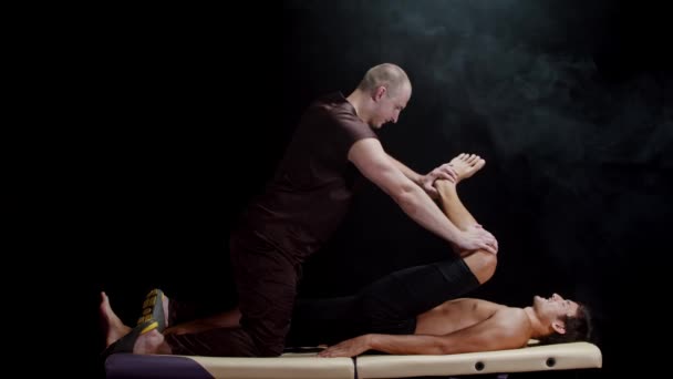 Chiropraktische Therapie im dunklen Studio - ein Therapeut arbeitet mit den Beinen seines Klienten - drückt das Bein in Richtung seines Körpers — Stockvideo