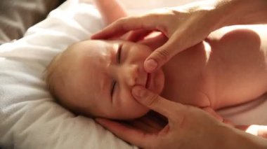 Yeni doğmuş bebek yatağında sırt üstü yatıyor ve ailesi yüzüne masaj yapıyor.