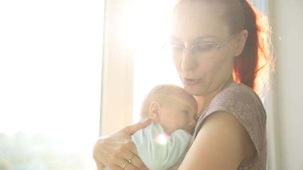 Mulher de gengibre em óculos segurando seu bebê em pé junto à janela — Vídeo de Stock