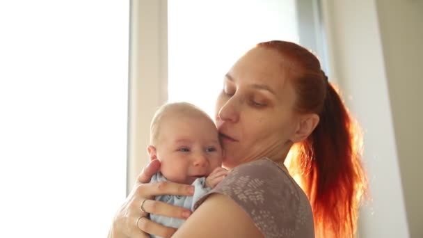 사랑하는 어머니가 어린 아기를 안고 창문 옆에 서 있는 아기를 쓰다듬고 있다 — 비디오