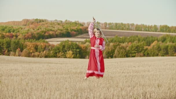 Joven hermosa mujer en vestido tradicional largo de pie en el campo y jugando con una espada larga — Vídeo de stock