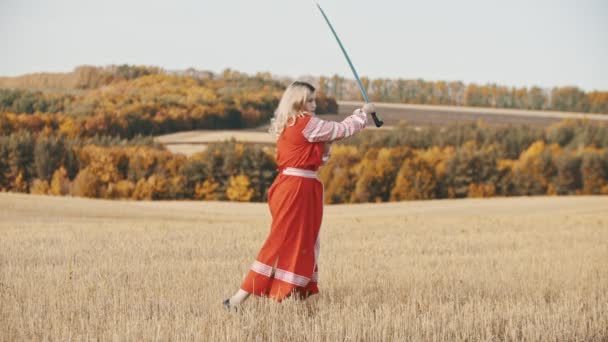 Jonge vrouw in lange traditionele jurk die op het veld staat en speelt met een lang zwaard — Stockvideo