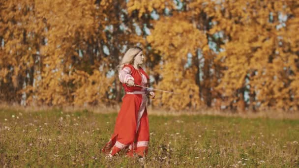 Kobieta w czerwonej narodowej sukience stojąca na polu i szermierka z mieczem — Wideo stockowe