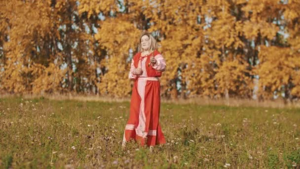 Blond vrouw in rode nationale jurk op het veld staan en schermen met een zwaard - gooit het op en vangen het — Stockvideo