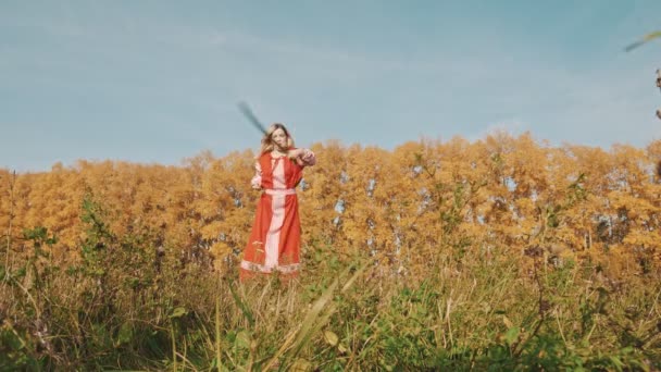 Mulher loira de vestido nacional vermelho em pé no campo e treinando sua esgrima com uma espada — Vídeo de Stock