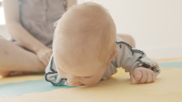 Маленький хлопчик лежить на животі на пастельному килимку і його мати сидить за ним — стокове відео