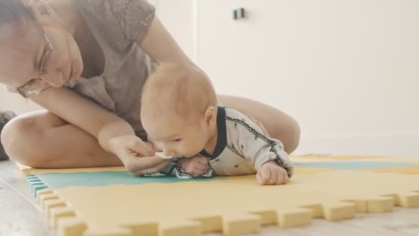 Little baby boy lying on his stomach on the pastel mat and his mother sitting behind him and wiping out his face with a cotton pad — Stock Video