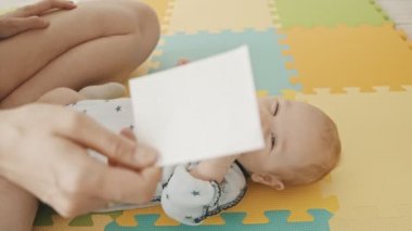 Anne bebeğiyle egzersiz yapıyor. Gözleriyle bir kağıt parçasını izlettirip takip ettiriyor.