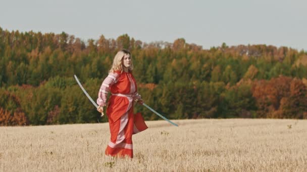 Vurige vrouw in rode jurk training op het veld - traint met twee zwaarden en roteert ze — Stockvideo