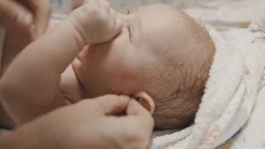 Küçük bir bebek yatakta yatıyor ve annesi kulak temizleyicisiyle kulaklarını temizliyor