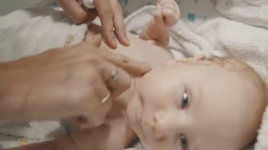 Küçük bir bebek yatakta yatıyor ve annesi boynuna ve vücuduna krem sürüyor.