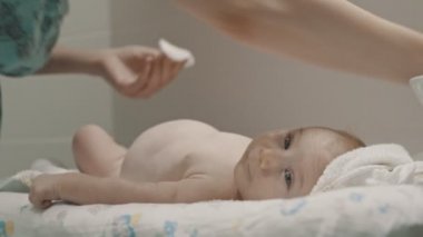 Küçük bir bebek yatakta yatıyor ve annesi bacağına krem sürüyor.