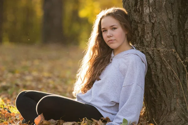 Fiatal nő tinédzser áll a fa mellett az őszi parkban — Stock Fotó