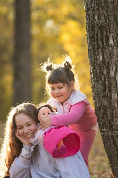 Dos hermanas sentadas en el suelo en el parque de otoño junto al árbol - una pequeña sosteniendo una muñeca — Foto de Stock