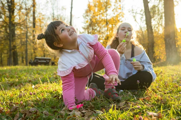 Dos hermanas en el parque de otoño: una niña sentada en el suelo y otra sentada a su lado y soplando burbujas de jabón — Foto de Stock