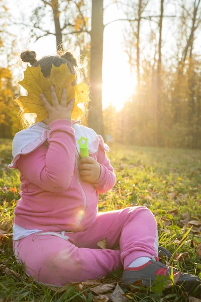 Una niña en traje rosa sentada en el suelo en el parque de otoño y cubriéndose la cara con una hoja grande — Foto de Stock