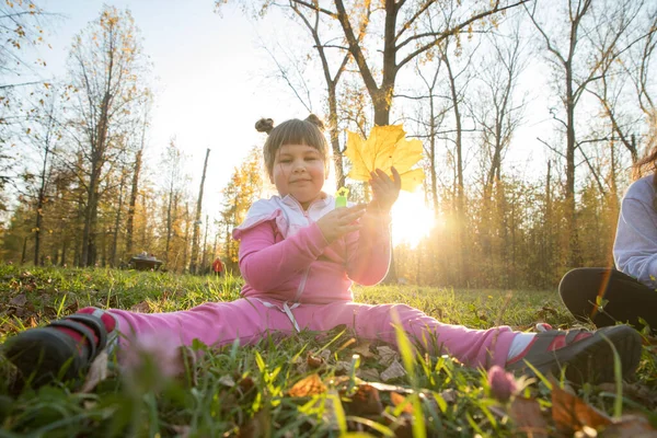 Una niña en traje rosa sentada en el suelo en el parque de otoño y sosteniendo una hoja grande — Foto de Stock