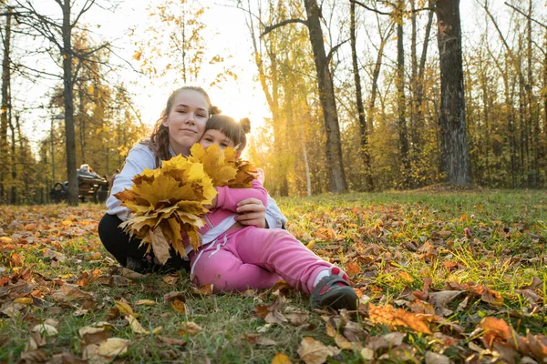 Dos hermanas pasan tiempo en el parque de otoño y recogen hojas anaranjadas, sentadas en el suelo y mostrándolas — Foto de Stock