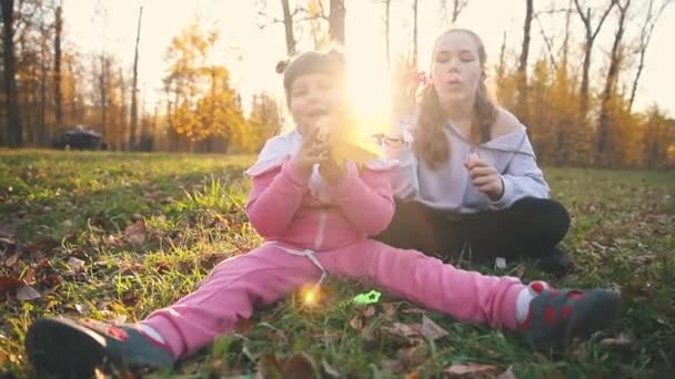 Due sorelle sedute a terra nel parco autunnale e una piccola che gioca con le foglie mentre un'altra soffia bolle di sapone — Video Stock