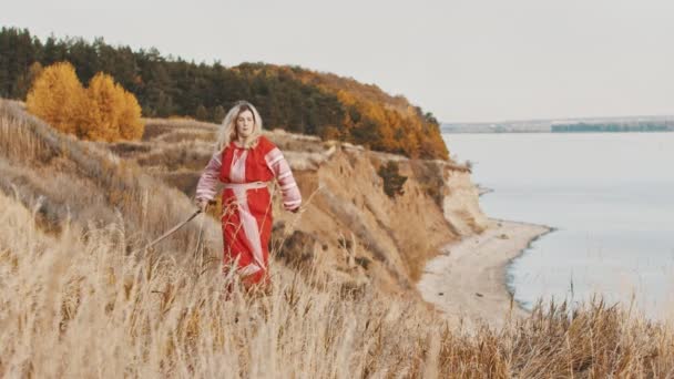 Kobieta w narodowej sukience stojąca na skraju wzgórza z dwoma mieczami i dzierżąca je. — Wideo stockowe