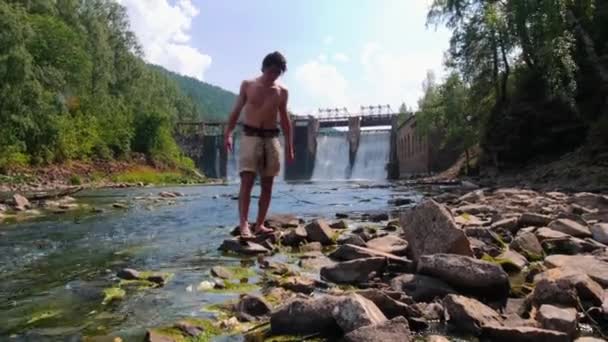 Vodní hráz v lese - mladý muž v kraťasech kráčející po skalách — Stock video