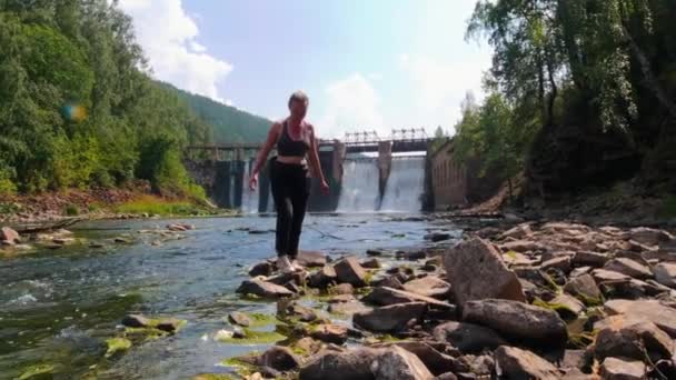 休暇中の若い女性-ストリーミング水の近くの岩の上を歩く-背景にある滝ダム — ストック動画