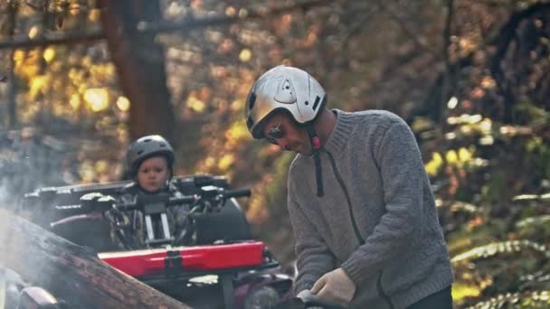 一个秋天的森林人在树林里打开一把链锯，他的儿子骑着一辆红色的四足摩托车等着他 — 图库视频影像