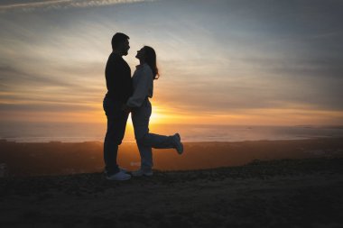 Sevgi dolu bir çiftin silueti bir tepenin üstünde, okyanusun üzerinde nefes kesen bir günbatımının tadını çıkarıyor.