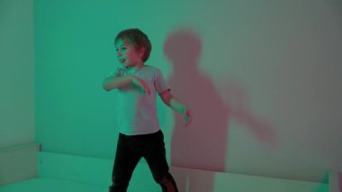 Enerjik çocuk yatakta dans eder ve oynar, canlı deniz mavisi ve pembe ışıklarla aydınlatılır, dinamik gölgeler bırakır.