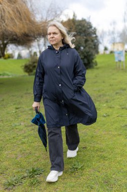 Yağmurluk giyen yaşlı bir kadın bulutlu bir günde katlanmış şemsiyeyle parkta yürüyor.