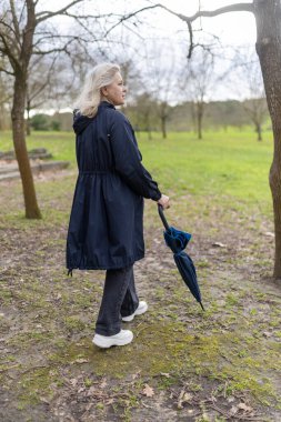 Üst düzey bir kadın, bulutlu bir günde elinde kapalı bir şemsiyeyle parkta huzurlu bir yürüyüşün tadını çıkarıyor, doğanın huzurunu kucaklıyor.