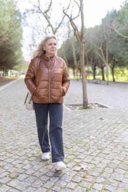 Yaşlı bir kadın kaldırımlı bir parkta sakin bir yürüyüş yapıyor, emeklilik sırasındaki doğanın huzurunu kucaklıyor.