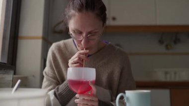 Kadın mutfak masasında oturmuş, soğuk, pembe bir içeceğin tadını çıkarıyor.