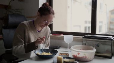 Genç bir kadın evinin penceresinin yanında makarna yiyor.