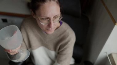 Gözlük takan ve günlük kıyafetler giyen kadın mutfaktaki bardaktan su içiyor.