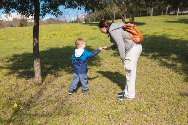 Anne güneşli bir günde yeşil bir parkta dururken oğluna bir şey veriyor.