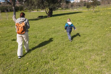 Sırt çantalı anne ve oğlu güneşli bir günde şehir parkında koşuşturuyorlar.