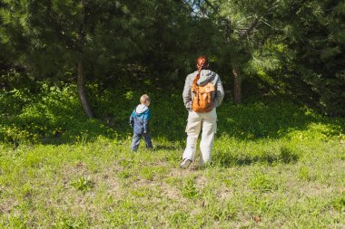 Deri çantalı anne ve oğlu güneşli bir günde yeşil ormanda yürüyüş yapıyor.