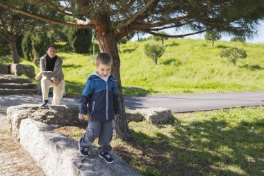 Genç bir çocuk parkta alçak bir taş duvar boyunca dikkatlice yürürken, dengesini ve koordinasyonunu geliştirirken, annesi yakınlarda onu izliyor.