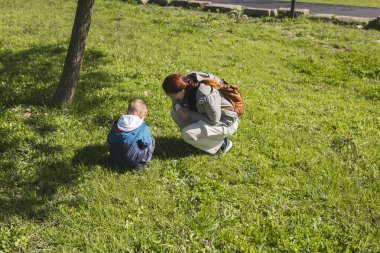 Anne ve oğul bir ağacın yakınındaki çayırda çömelmiş, doğayı keşfediyor ve yerde bir şey gözlemliyorlar.