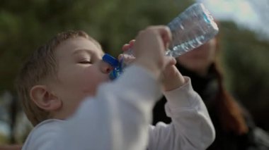 Parktaki plastik bir şişeden su içen genç bir çocuk, annesi onu izliyor.