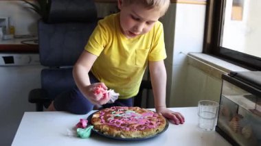 Genç çocuk bir ev mutfağında pembe kremayla pasta süslüyor.