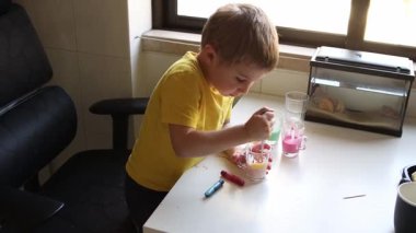 Genç bir çocuk renkli sıvılarla deney yapıyor, onları beyaz bir masada bir bardakta karıştırıyor.