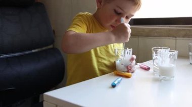 Genç bir çocuk beyaz bir masada renkli bir macunu bir bardak süte karıştırıyor.