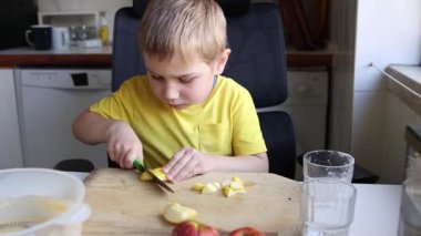 Sarışın çocuk mutfaktaki tahta bir tahtanın üzerinde bıçakla elmaları küçük parçalara ayırmayı öğreniyor.