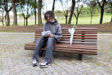 Kıvırcık saçlı genç adam, yanında kapalı şeffaf bir şemsiyeyle parktaki ahşap bir bankta oturan not defterine yazı yazıyor.